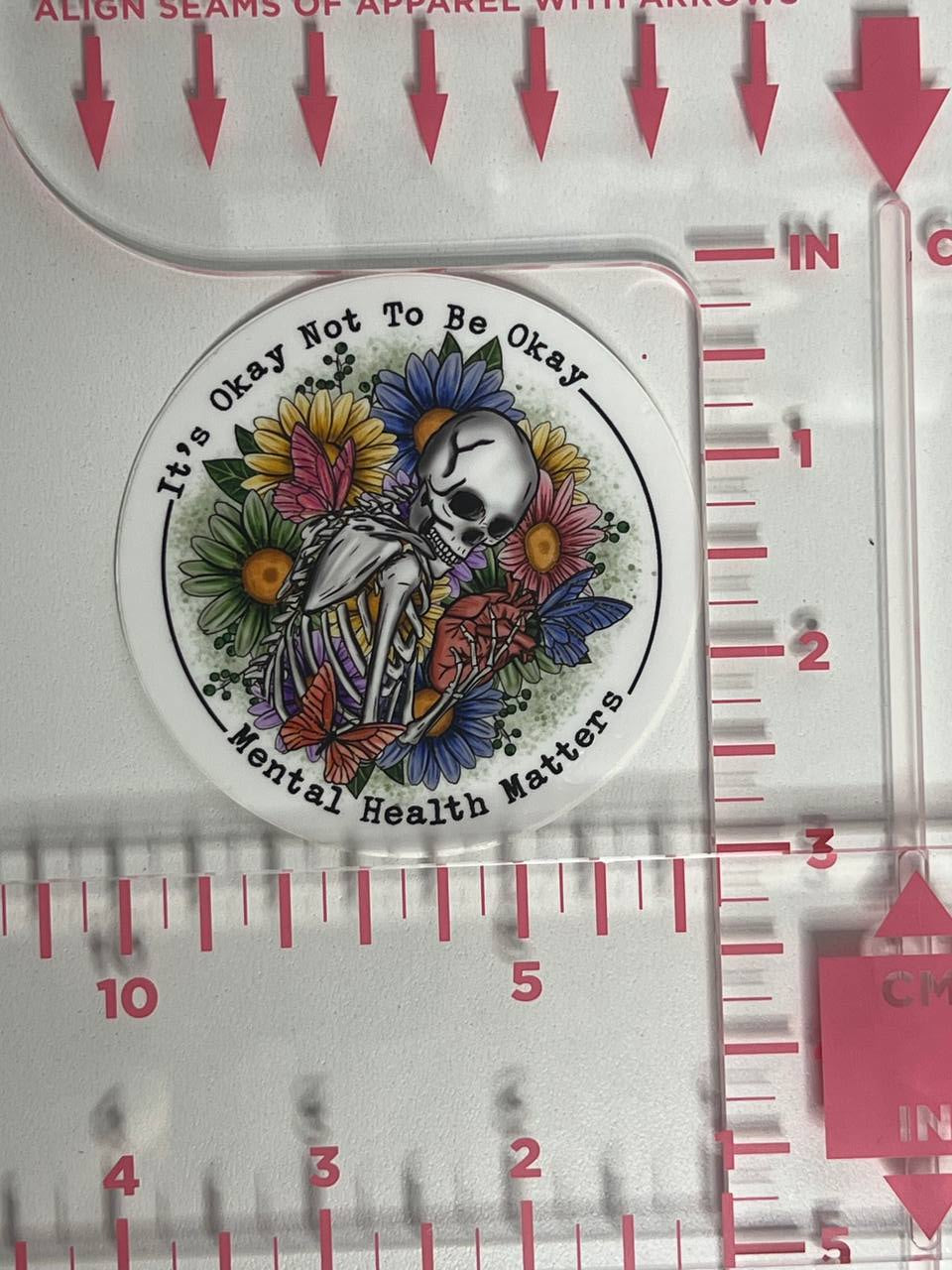Circular sticker with a skull and flowers design on a ruler background