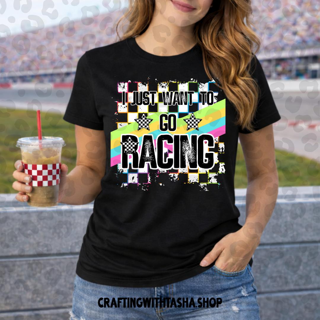 Person wearing a black t-shirt with 'I just want to go racing' design, holding a drink at a race track.