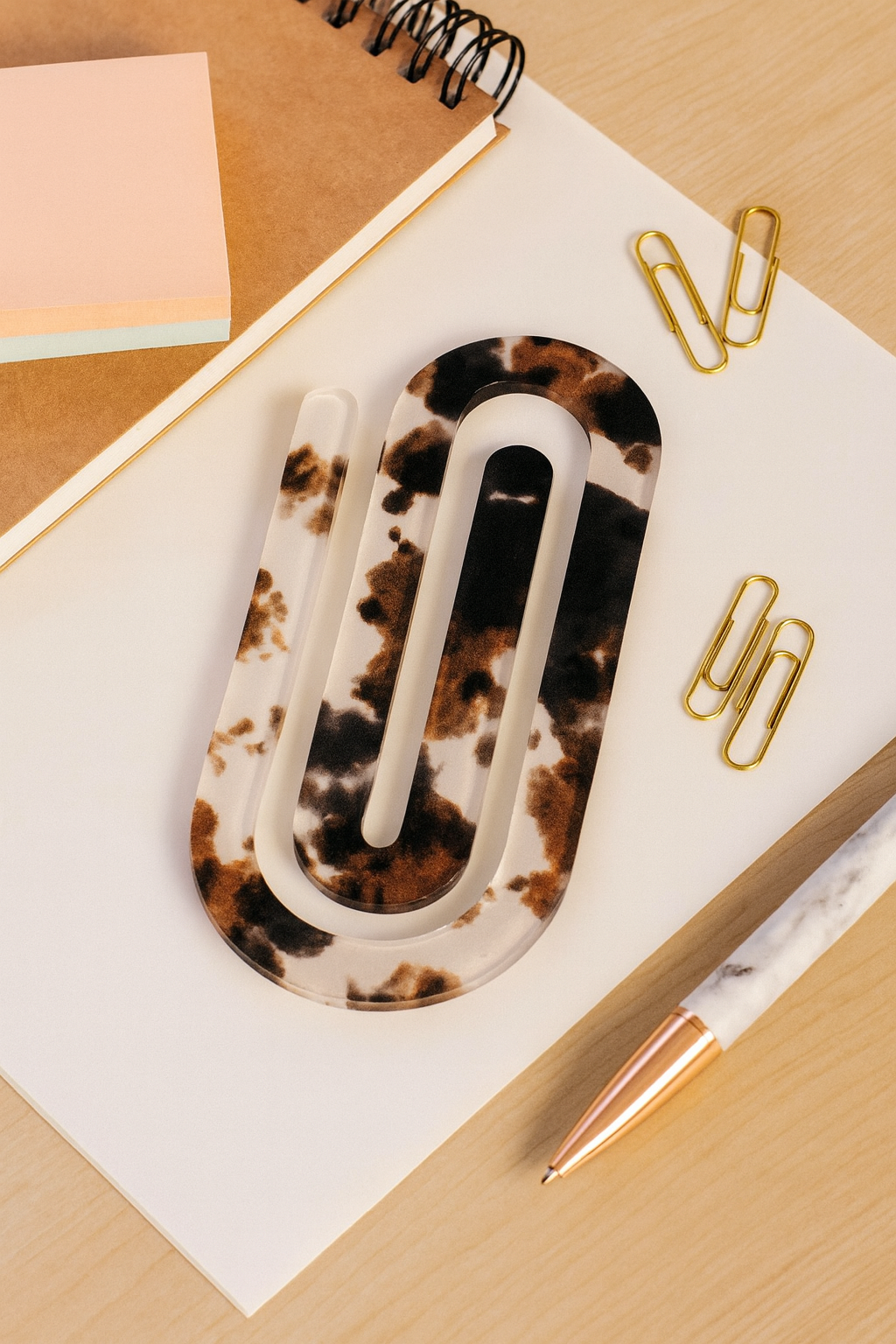 Cow print acrylic jumbo paper clip on a light wood desk with notebook, pen, and pastel stationery in soft daylight.
